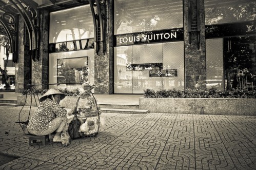 Sự tương phản thường gặp: Một người phụ nữ bán bánh ngoài cửa hàng của thương hiệu túi xách thời trang cao cấp Louis Vuitton.